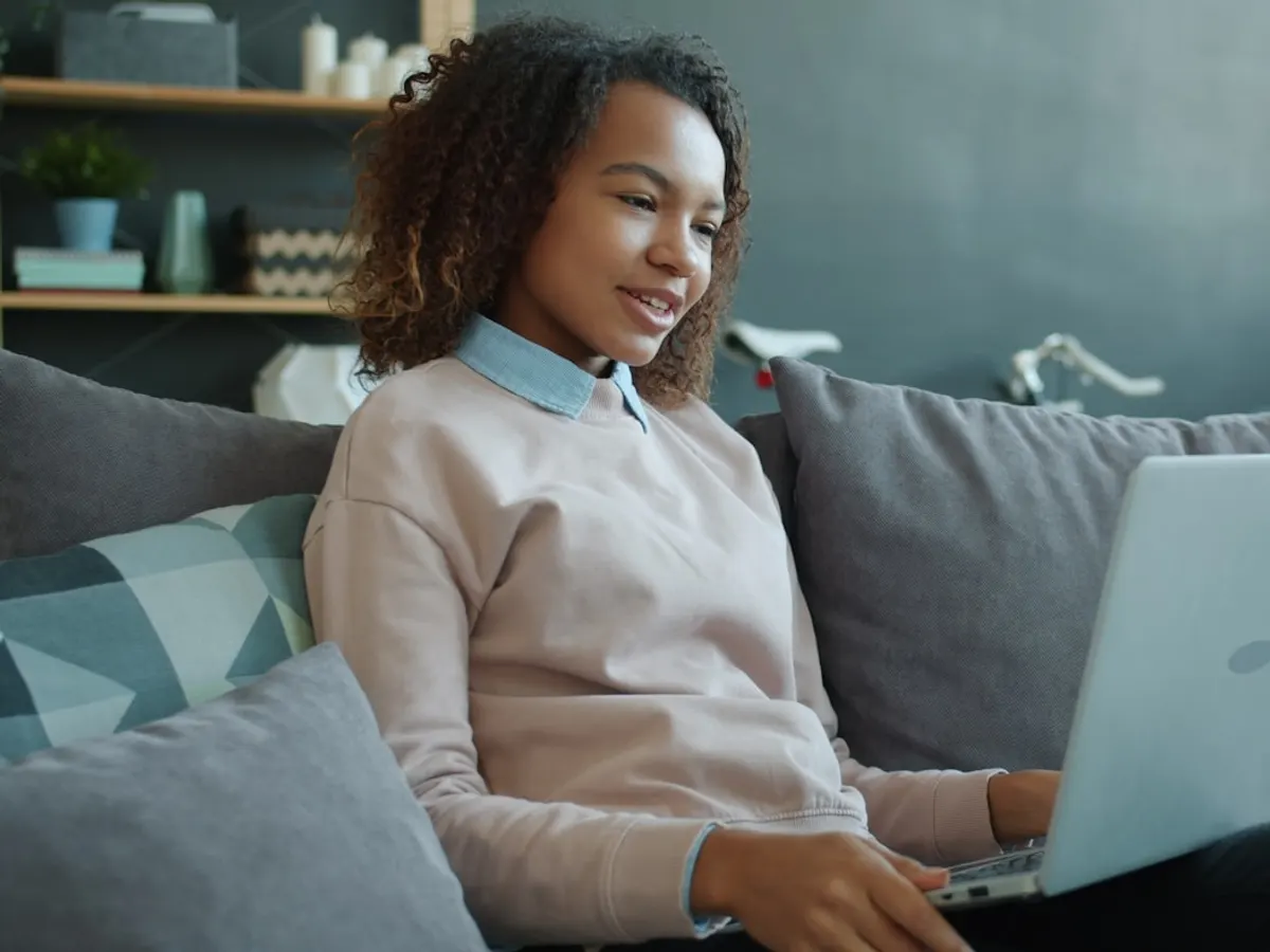 Woman relaxing on her couch at home, smiling while using a laptop for an online healthcare visit